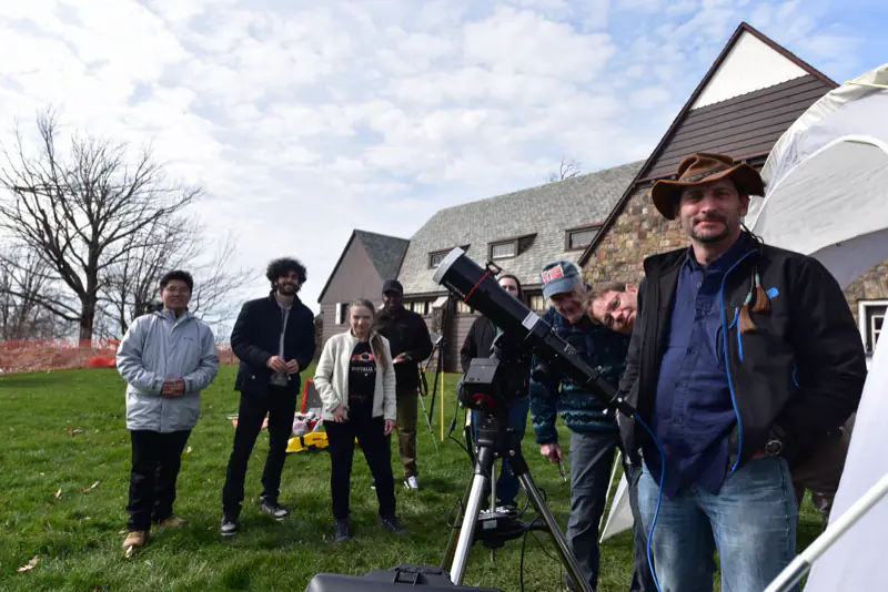 Eclipse observation team members on a lawn behind a telescope on a tripod, with a stone lodge and a white tent in the background.