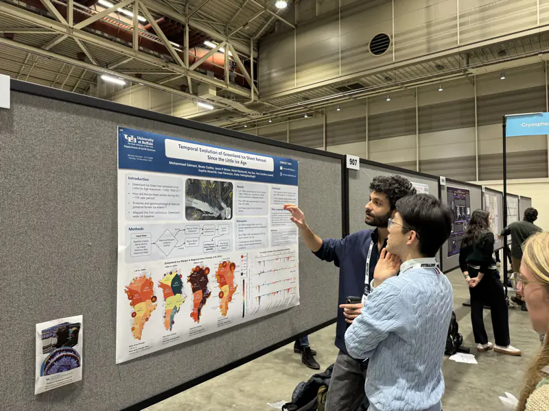 (a) Mohammad Salmani pointing at his poster, which shows four colored maps of Greenland, while a visitor looks on.