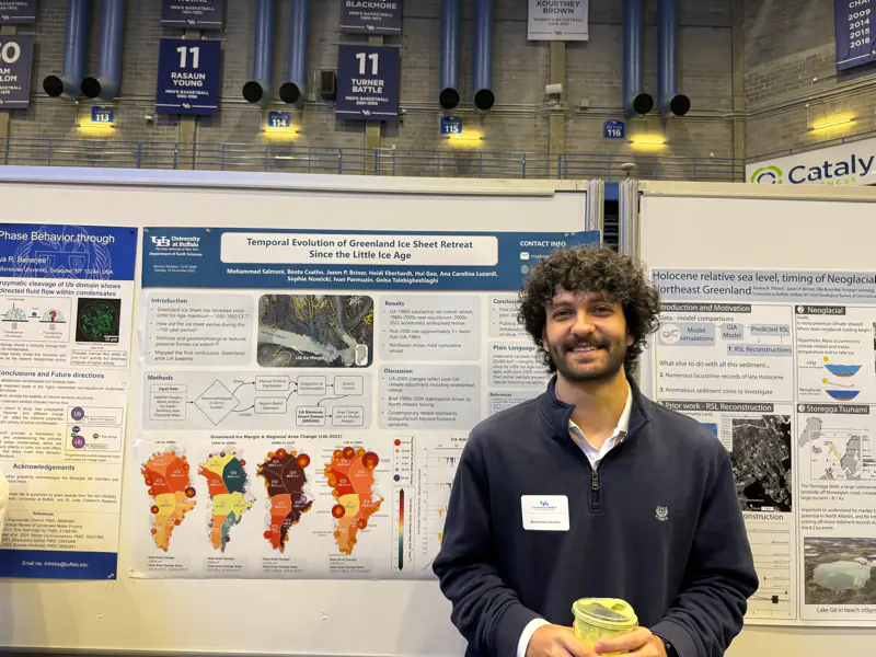 (b) Mohammad Salmani, holding a cup, in front of his poster “Temporal Evolution of Greenland Ice Sheet Retreat Since the Little Ice Age” in a gym set up for the poster session.