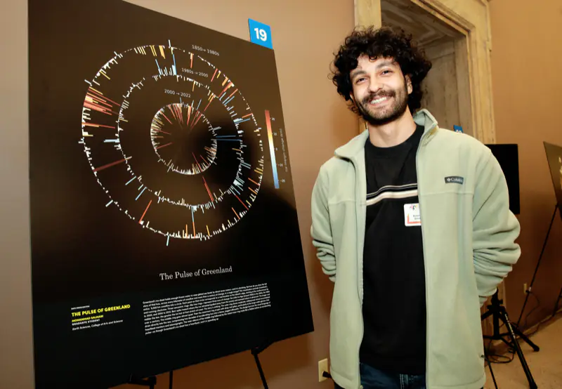 Mohammad Salmani smiling beside “The Pulse of Greenland”, a dark print of concentric radial bar rings for the periods 1850 to the 1980s, the 1980s to 2000, and 2000 to 2022.